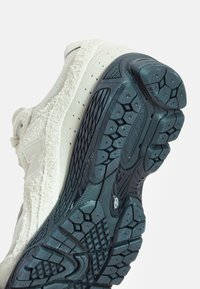 Fehér textil sportcipő, strukturált felső résszel és logóval, masszív fekete gumitalppal, amely háromszög alakú bordázással rendelkezik.