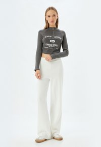 Grey ribbed crop top with a high zip neck and white print, paired with white wide-leg pants and grey shoes with a light sole.