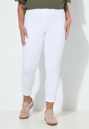 Person trägt weiße kurze Leggings und hellbeige Turnschuhe, steht vor einem schlichten hellen Hintergrund.