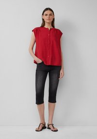 Czerwona bluzka na krótki rękaw z zapięciem na guziki i plisowanymi detalami, w połączeniu z ciemnymi, ściętymi dżinsami i czarnymi klapkami.