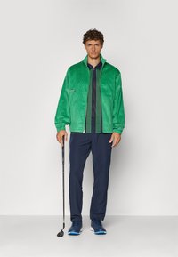 Grønn cordfløyelsjakke med høy krage, marineblå polo under, marineblå bukser og sko med blå detaljer. Holder en golfklubbe.