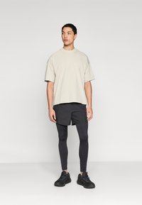 Μπεζ φαρδιά μπλούζα με κοντά μανίκια, συνδυασμένη με μαύρο αθλητικό σορτς και μαύρα κολάν, φορεμένη με μαύρα αθλητικά παπούτσια.