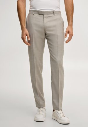 Mann trägt beige Slim-Fit-Anzughose und weiße Sneakers, steht vor hellem, einfarbigem Hintergrund.