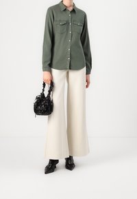 Chemise boutonnée vert olive avec deux poches poitrine, associée à un pantalon crème à jambes larges, des chaussures noires à bouts pointus, et un petit sac à main noir à motif.