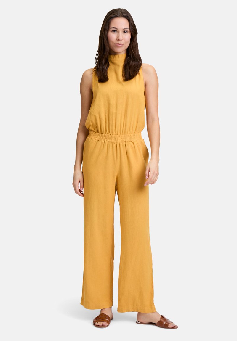 Geel, mouwloze jumpsuit met een hoge halslijn en elastische taille; wijd uitlopende broekspijpen. Gecombineerd met bruine instapsandalen.