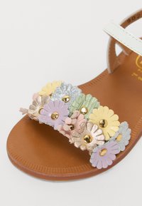 Sandale plate avec une semelle marron, une sangle blanche et une bande décorative de fleurs texturées multicolores avec des accents dorés.