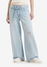 Lys blå vide jeans laget av denim, med høy midje, slitte detaljer og et bundet belte. Stylet med hvite sneakers.