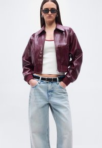 Γυναίκα που φοράει σκούρο κόκκινο δερμάτινο μπουφάν, λευκό τοπ, ανοιχτόχρωμο μπλε τζιν, μαύρη ζώνη, ασημένια κρίκους σκουλαρίκια και μαύρα ορθογώνια γυαλιά ηλίου.