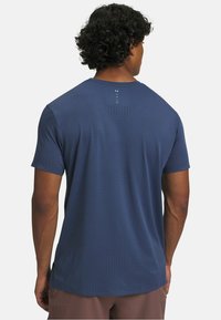 Homme aux cheveux bouclés portant un t-shirt de sport bleu marine à manches courtes perforé, vu de dos sur un fond blanc.