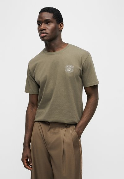 Νέος άνδρας που φοράει ελιάς πράσινο κοντομάνικο μπλουζάκι με μικρό λευκό κεντημένο λογότυπο στο στήθος και καφέ παντελόνι, στέκεται με το ένα χέρι στην τσέπη.