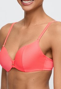 Korallinpunainen bikiniyläosa, jossa on ribbineulosta, kaarituet, säädettävät hihnat ja kullanvärinen solki keskellä edessä.