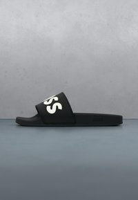 Sandale noire à bride large avec de grandes lettres blanches "SS" et nom de marque embossé sur le côté de la semelle.