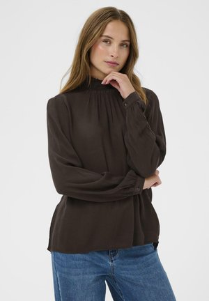 Bluză lungă, din material maro, cu mâneci lungi, guler înalt cu volane și manșete cu nasturi. Textură moale, design pliat și croială lejeră, completată de blugi albaștri.