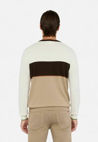 Man gezien vanuit de achterkant, gekleed in een sweater met lange mouwen met witte, donkerbruine en beige kleurvlakken, gecombineerd met beige broek.