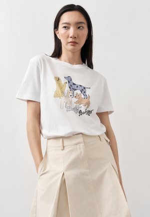 Młoda kobieta z długimi ciemnymi włosami, ubrana w biały t-shirt z kolorowymi ilustracjami psów i beżową spódnicę z wysokim stanem, ręce w kieszeniach.