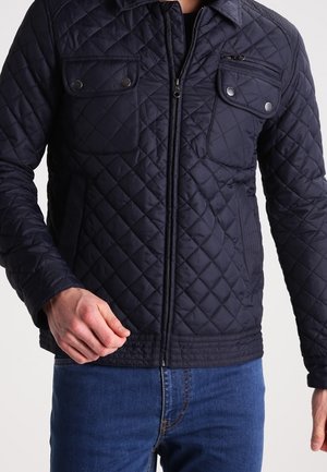 Navy quilted jacket with a zip closure, two chest pockets, and textured shoulder accents. Smooth fabric with a fitted design.