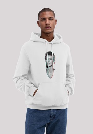 Young man with short hair wearing a white hoodie featuring a black-and-white portrait of a person with a cross necklace, paired with blue jeans.