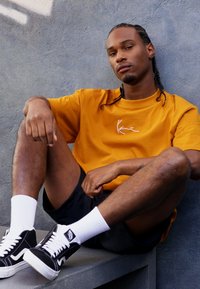 Tricou din bumbac portocalie cu logo alb, șorturi negre și pantofi sport Vans negri cu accente albe, așezat lângă un perete gri.