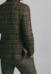 Blazer a quadre marrone con un design strutturato, tessuto testurizzato e chiusura a un bottone. Presenta aperture laterali e bottoni sulle maniche.