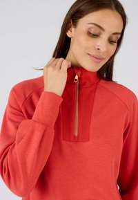 Rode sweatshirt met hoge kraag, geribbeld textuur bij de hals en een gouden ritsdetail. Zacht materiaal met lange mouwen en een ontspannen pasvorm.