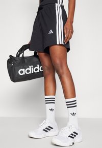 Shorts de sport noirs avec des rayures blanches, chaussettes blanches avec des rayures et un logo noirs, et chaussures blanches avec des accents noirs. Portant un sac noir avec un logo.
