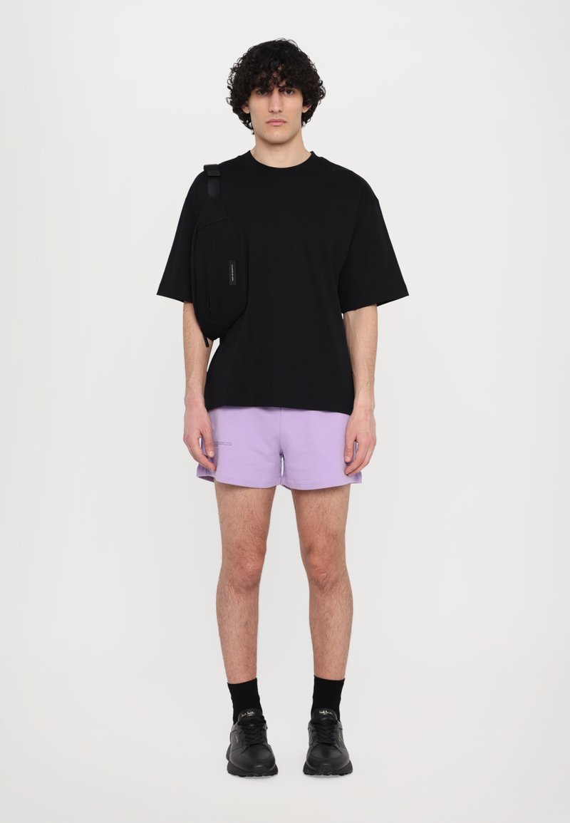 Camiseta negra de manga corta combinada con pantalones cortos de color púrpura claro. Bolso bandolera negro en el lado izquierdo, zapatillas negras y calcetines negros.