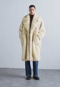 Šviesiai rudos dirbtinės kailio paltas su plačia apykakle ir giliomis kišenėmis, derinamas su tiesiomis mėlynomis džinsomis ir juodais batais, neutraliame fone.