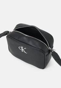 Borsa a tracolla in pelle nera con logo argentato, forma rettangolare, texture liscia, chiusura con zip e tracolla regolabile. Interno in tessuto nero.