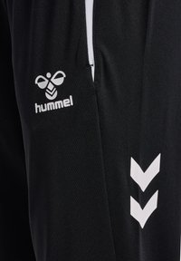Tissu noir avec logo blanc de Hummel et deux bandes chevrons vers le bas, gros plan sur un détail de vêtement de sport.