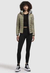 Gewatteerde olijfgroene jas met een grijze capuchon, zwarte leggings en zwarte hoge sneakers. Gladde textuur met een ritssluiting aan de voorkant en een getailleerd ontwerp.