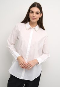 Camicia bianca con collo arrotondato decorato con accenti di perle. Maniche lunghe e orlo curvo, realizzata in tessuto leggero.