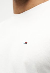 Κοντινό πλάνο στο πάνω μέρος του στήθους ενός ατόμου που φοράει μια απλή λευκή μπλούζα με στρογγυλή λαιμόκοψη και ένα μικρό ορθογώνιο λογότυπο με μπλε σκούρες, λευκές και κόκκινες ρίγες.