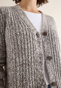 Pletený cardigan v šedých a krémových odstínech, s výstřihem do V, texturovaným vzorem a tmavě hnědými knoflíky vpředu.