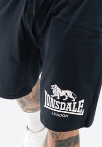 Mustat puuvillahousut, joissa on valkoinen Lonsdale-logo ja leijonagraafikka vasemmalla lahkeella. Suunnittelussa on joustava vyötärönauha, jossa on nyöri.