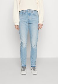 Világoskék, karcsú szabású farmernadrág, amely denim anyagból készült. Öt zsebe van, finoman kopott területekkel és enyhe elasztikus anyaggal. Szürke sportcipővel párosítva.
