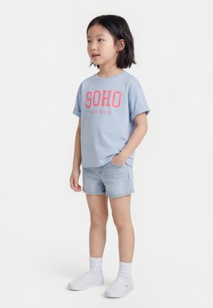 Młoda dziewczyna stojąca bokiem, ubrana w jasnoniebieską koszulkę z napisem „SOHO New York”, jeansowe szorty, białe skarpetki i białe buty wsuwane.