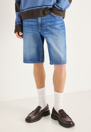 Jeansshorts in Hellblau mit geradem Schnitt, ausgefranstem Saum und klassischem Fünf-Taschen-Design, kombiniert mit dunkelbraunen Loafers und weißen Socken.
