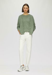 Cardigan verde a maglia con bottoni, indossato sopra un maglione abbinato, abbinato a pantaloni bianchi dritti e sneakers grigie camouflage.