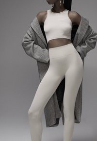 Donna che indossa un crop top senza maniche beige e leggings, con un cappotto grigio oversize sceso sulle spalle su uno sfondo grigio uniforme.
