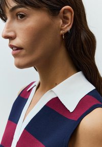 Haut sans manches rayé avec un col blanc ; les couleurs incluent le marine, le bordeaux et la crème. Le mannequin porte des boucles d'oreilles en or et en argent.