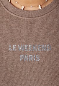 Bruine sweatshirt met geribbelde ronde hals. Heeft zilveren glittertekst "LE WEEKEND PARIS" in vetgedrukt, gecentreerd op de voorkant. Gladde stoftextuur.