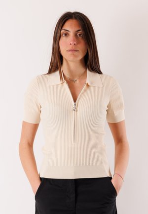 Donna con capelli castani lunghi che indossa una maglia crema a maniche corte a coste con una cerniera frontale e pantaloni neri, in piedi con le mani nelle tasche.