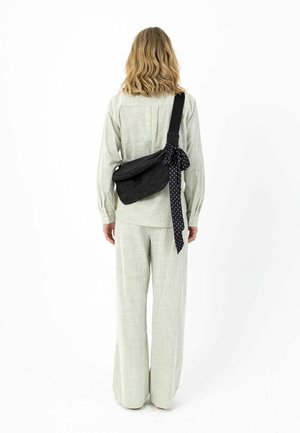 Osoba odjevena u svijetlu kariranu košulju i hlače, s crnom torbom na rame s točkastim šaljem zavezanim na remenu, okrenuta leđima na bijeloj pozadini.