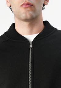 Chaqueta negra con cremallera y cuello y puños acanalados, con una cremallera plateada y una textura de tela suave. Vista recortada desde el pecho hacia arriba.