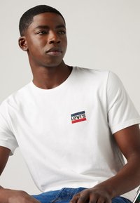 Balts kokvilnas T-krekls ar mazu sarkanu, tumši zilu un baltu Levi's logotipu kreisajā pusē. Vienkāršs apļa kakla dizains, īsas piedurknes.