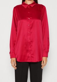 Czerwona satynowa koszula z wywiniętym kołnierzykiem, długimi rękawami i zapięciem na guziki. Gładka faktura z lekkim połyskiem i stylowymi detalami przy mankietach.