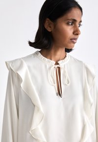 Balta blūze ar volāniem uz pleciem, noapaļotu kakla izgriezumu un sasienamu aizdari ar metāla galiem. Gluda, gaisīga auduma. 