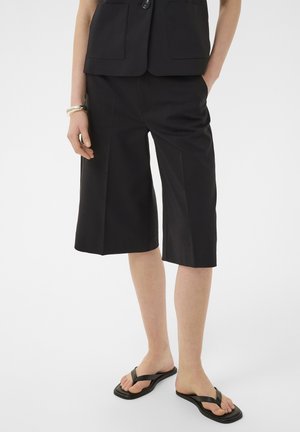 Személy fekete térdig érő, bő szárú rövidnadrágot, fekete ujjatlan mellényt, fekete flip-flop papucsot visel, egyik kezét zsebében tartja, csuklóján karkötők vannak.