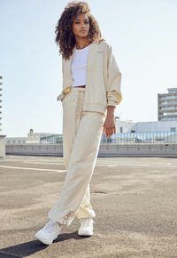 Beige Trainingsanzug in lockerer Passform, bestehend aus einer Jacke mit Reißverschluss und einer Hose mit elastischem Bund. Weiße Sneakers mit strukturiertem Design.