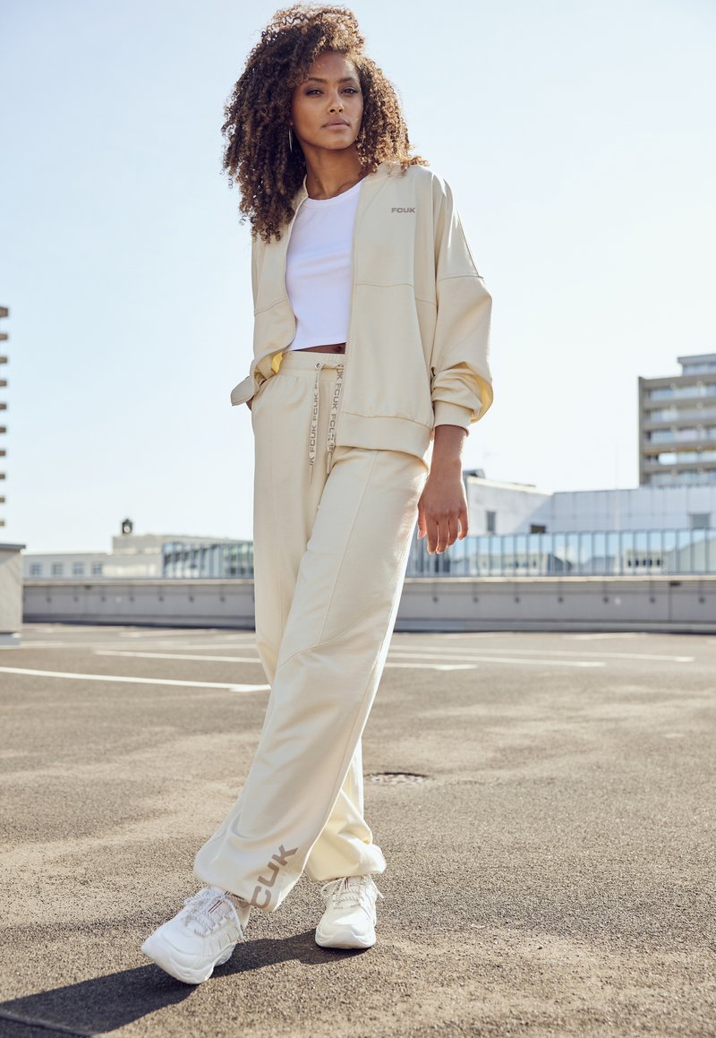 Beige Trainingsanzug in lockerer Passform, bestehend aus einer Jacke mit Reißverschluss und einer Hose mit elastischem Bund. Weiße Sneakers mit strukturiertem Design.